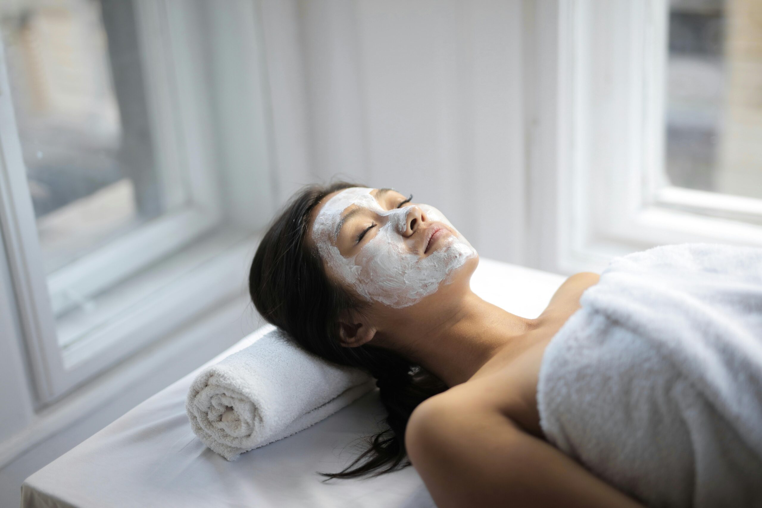 Young Asian woman on towel and with mask on face lying on covered table in light spa salon